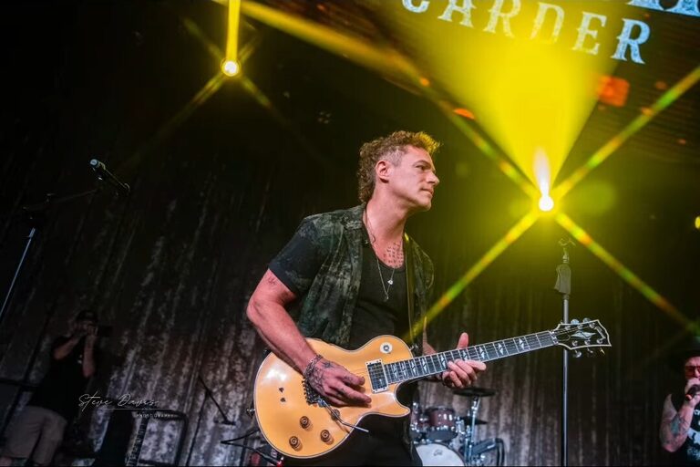Guitarist playing a gold electric guitar beneath yellow stage beams.