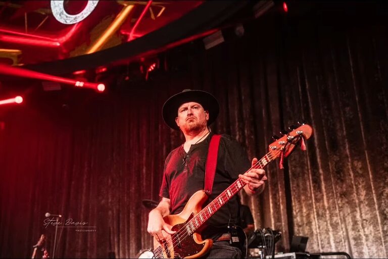Bassist in a hat performing under red stage lights.